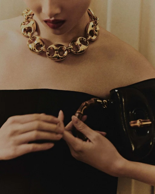 Elegant woman showcasing a gold chain necklace and a black patent leather handbag, highlighted by indoor studio lighting.