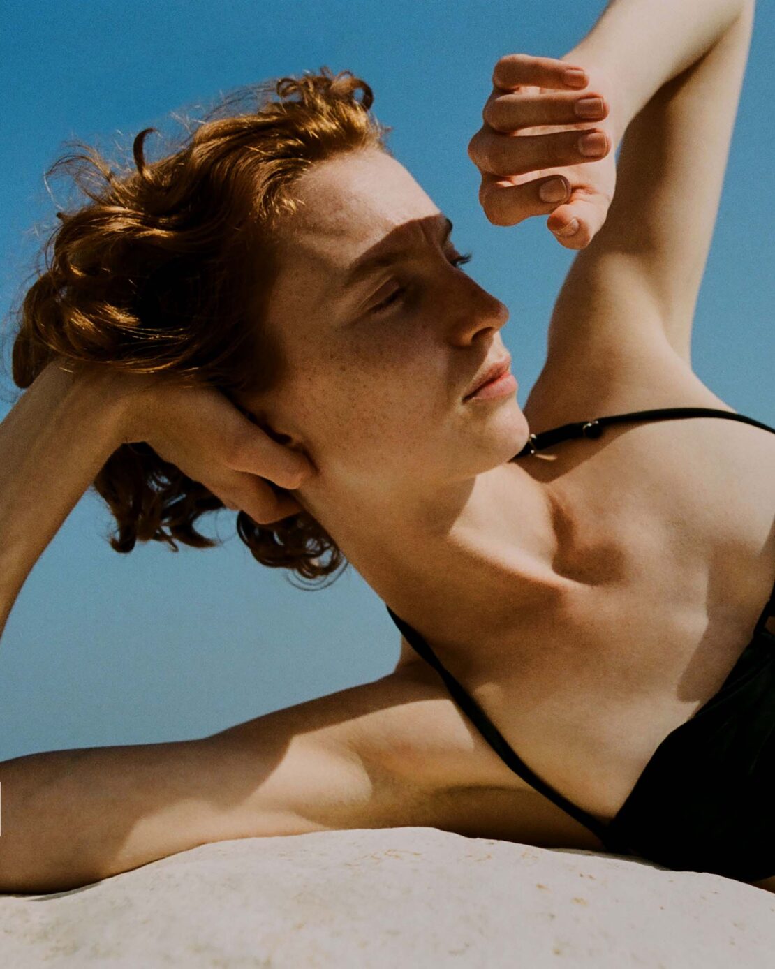 Close-up of a red-haired woman lying in the sand, wearing a black swimsuit, with her hands behind her head under a bright blue sky.