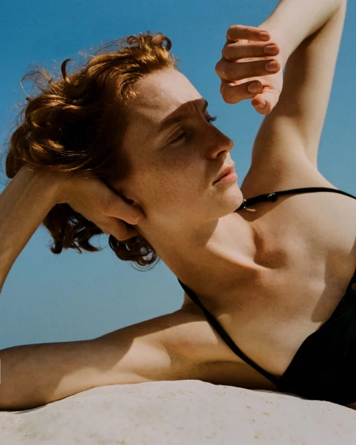 Close-up of a red-haired woman lying in the sand, wearing a black swimsuit, with her hands behind her head under a bright blue sky.