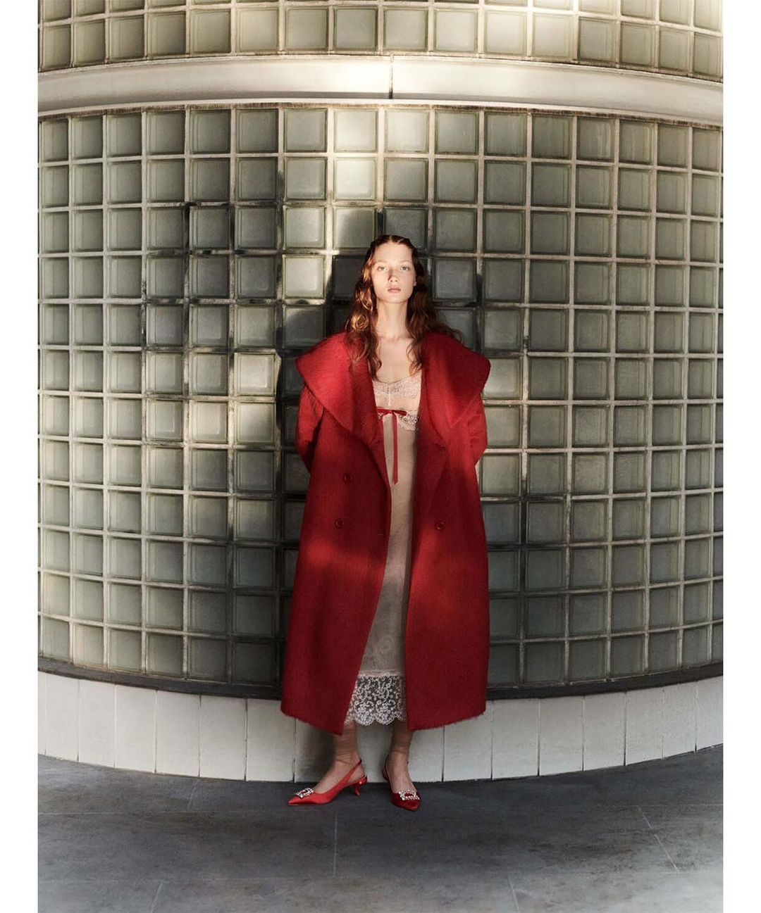 An adult female is posing outdoors in a red coat and slingback heels against a patterned wall on a cloudy day.