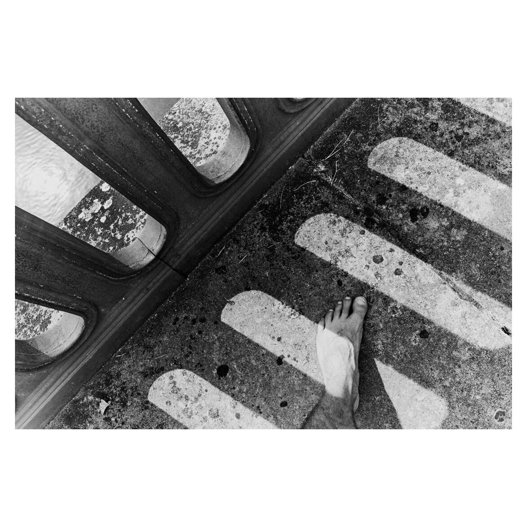 Black and white photograph shows a bare foot on a concrete surface near a metal grate, capturing texture and contrast.