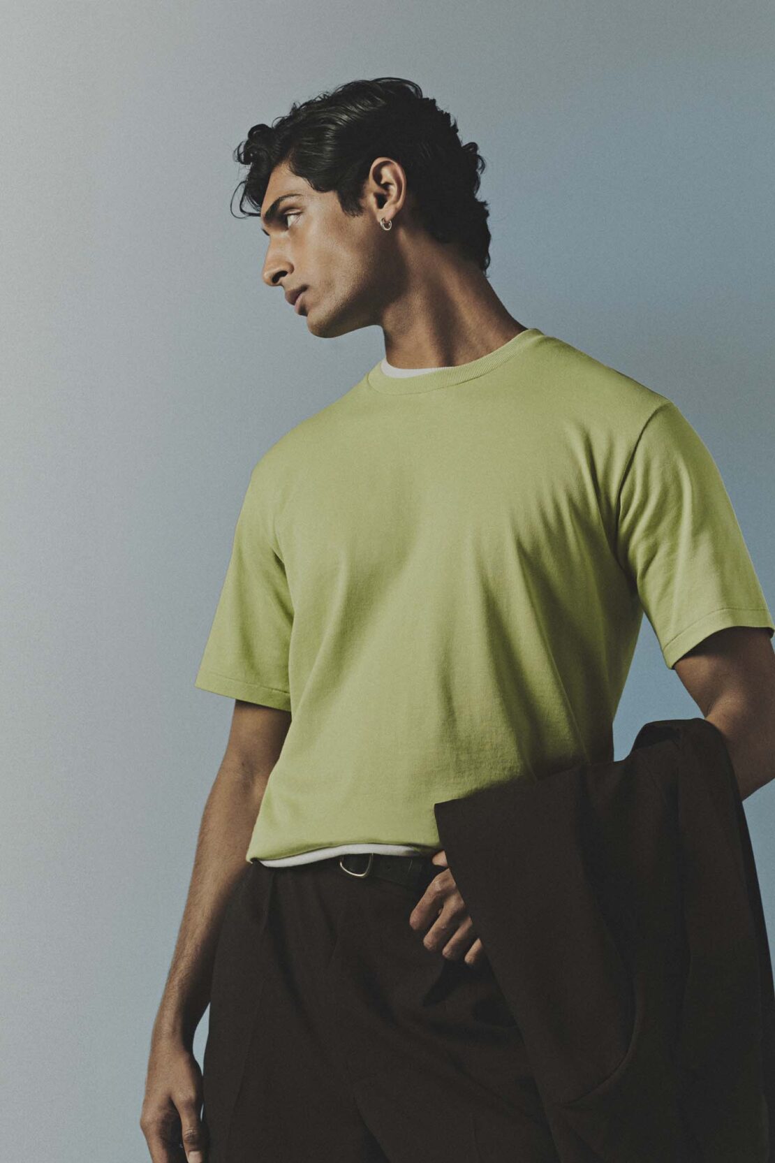 Portrait of a man in a green t-shirt holding a dark jacket against a light blue gradient backdrop.