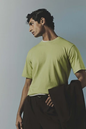 Portrait of a man in a green t-shirt holding a dark jacket against a light blue gradient backdrop.