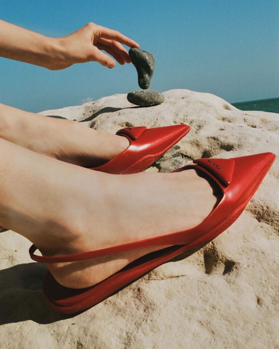 Red fashion flats, feet, and hands shown on a rock formation with blue sky and ocean background; styled product fashion, outdoor setting.