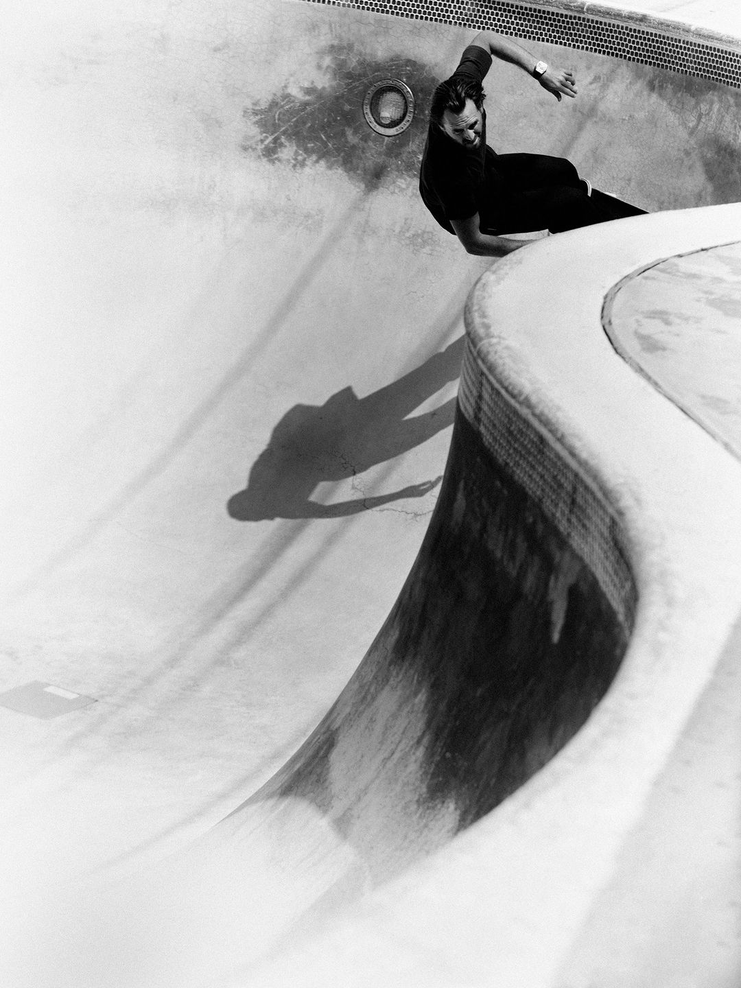 Monochrome photo of a skateboarder riding the curved wall of an empty pool, casting a shadow on the worn concrete surface.