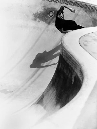 Monochrome photo of a skateboarder riding the curved wall of an empty pool, casting a shadow on the worn concrete surface.