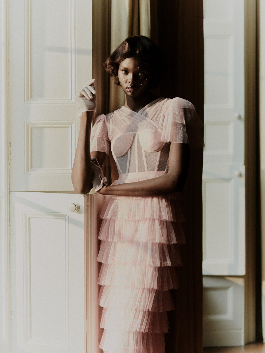 Elegant Black woman in a layered pink gown poses by white doors, adorned with long white gloves, in a softly lit indoor fashion portrait.