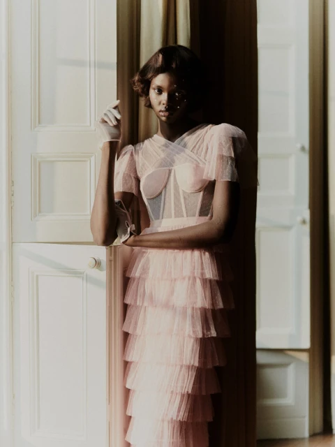 Elegant Black woman in a layered pink gown poses by white doors, adorned with long white gloves, in a softly lit indoor fashion portrait.