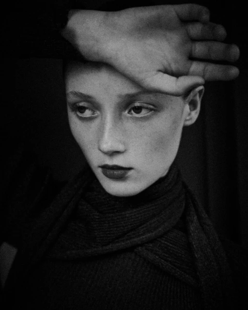 Monochrome portrait of a fair-skinned young adult with dark lipstick and a hand resting on their forehead, set against a dark background.