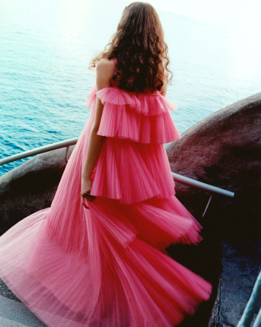 Woman in a pink, layered gown stands on a stone pathway, looking out over the ocean, styled for a fashion editorial.