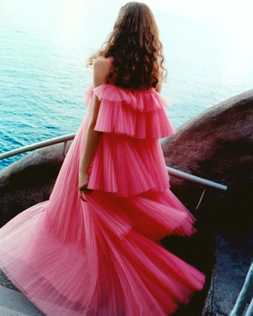 Woman in a pink, layered gown stands on a stone pathway, looking out over the ocean, styled for a fashion editorial.  