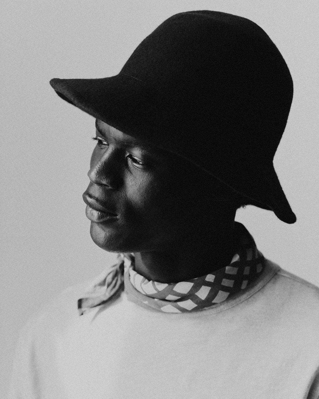 Portrait of a Black man wearing a wide-brimmed black hat and patterned neck scarf against a neutral background.