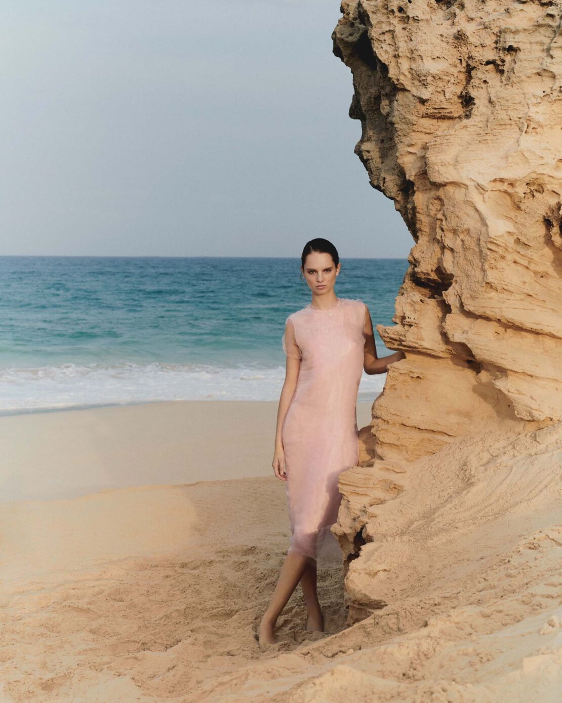 Woman in pale pink dress standing on sandy beach beside a rocky outcrop with ocean view