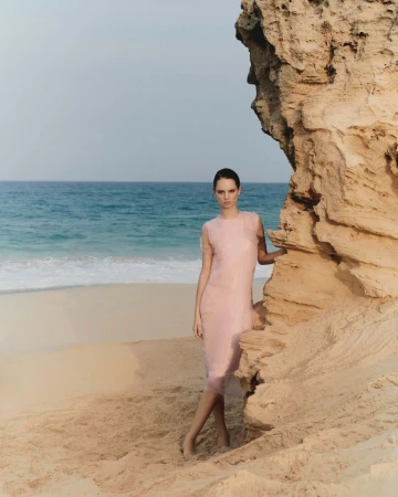 Woman in pale pink dress standing on sandy beach beside a rocky outcrop with ocean view