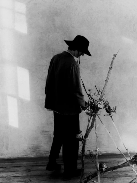 Monochromatic portrait of a man in a wide-brimmed hat next to a bare floral arrangement against a textured wall with window light.
