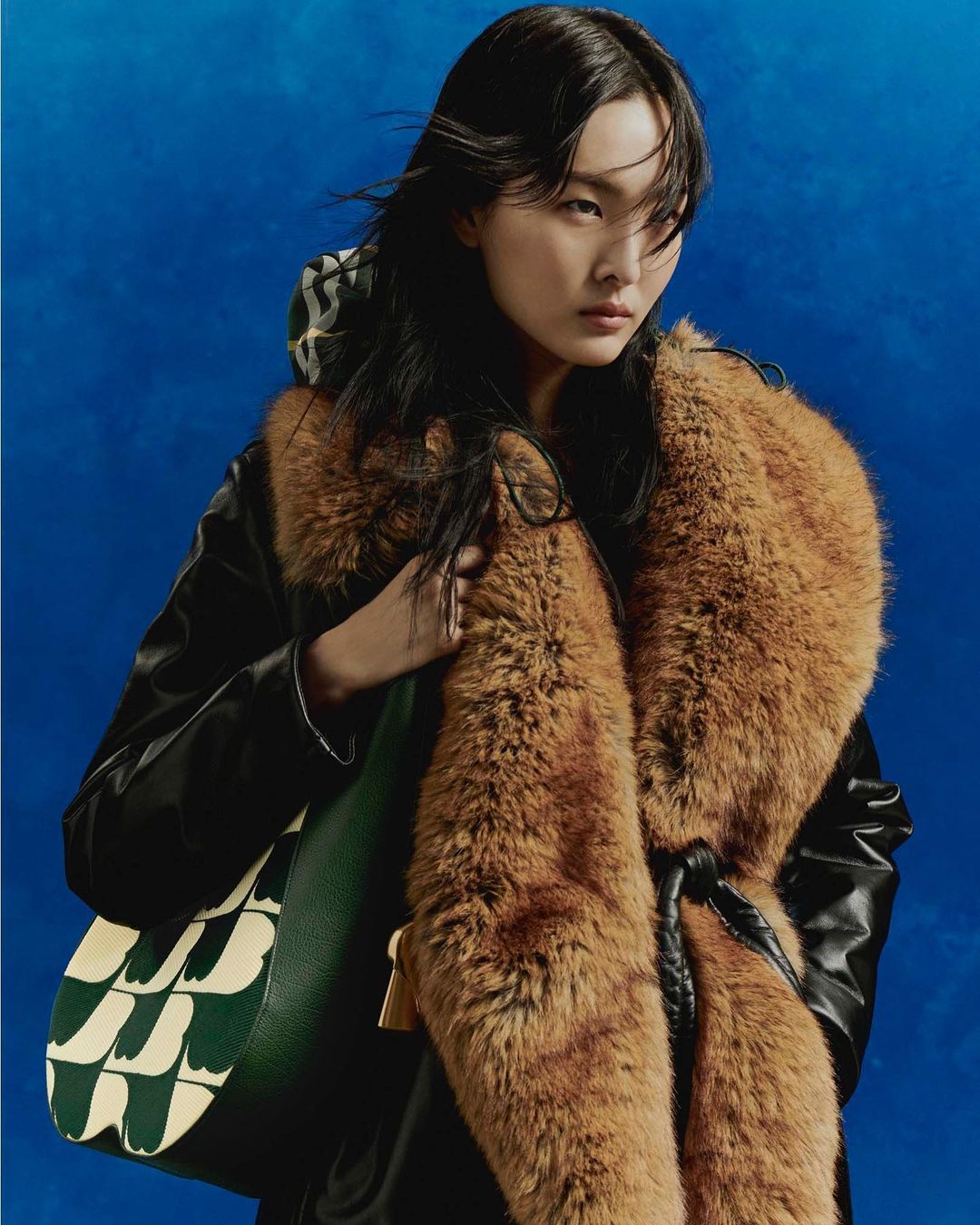 East Asian woman wearing a black jacket with a large brown faux-fur collar, holding a patterned green bag against a textured blue backdrop