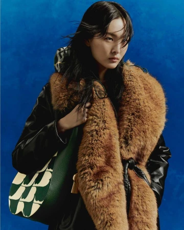 East Asian woman wearing a black jacket with a large brown faux-fur collar, holding a patterned green bag against a textured blue backdrop