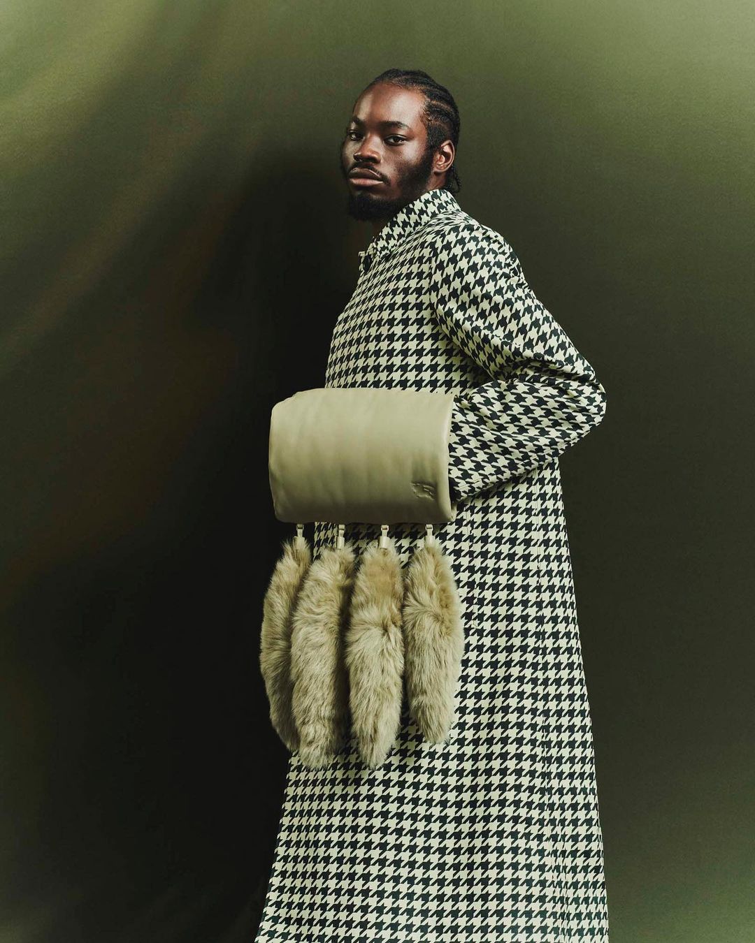 Black man in houndstooth coat holding a textured cylindrical handbag with fur charms against a dark olive backdrop
