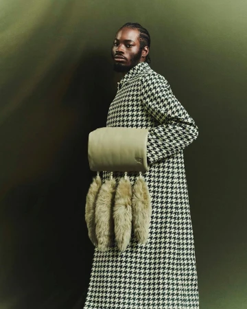Black man in houndstooth coat holding a textured cylindrical handbag with fur charms against a dark olive backdrop