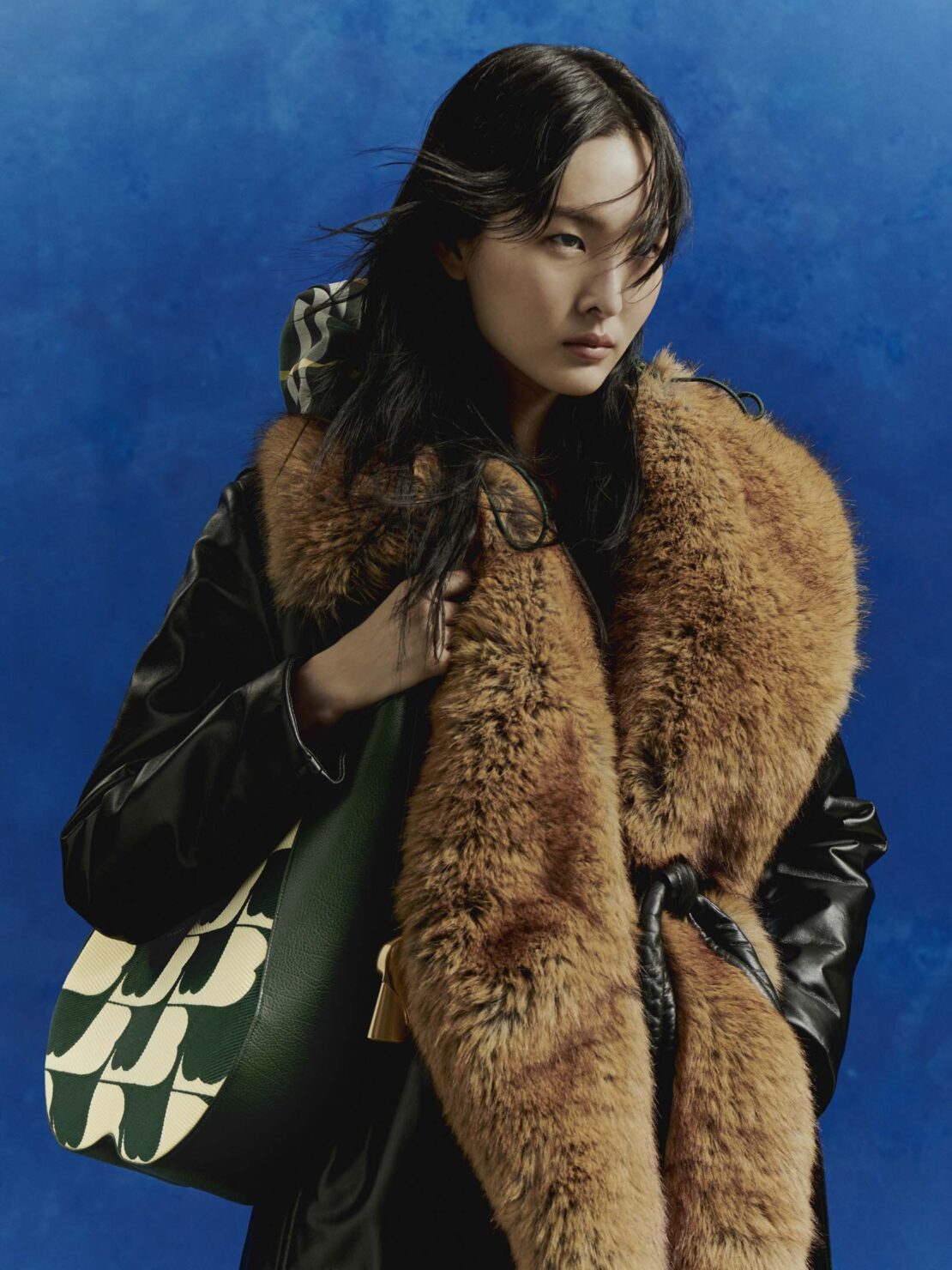 East Asian woman in a black coat with a faux fur collar holding a green patterned shoulder bag against a blue backdrop.