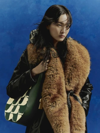 East Asian woman in a black coat with a faux fur collar holding a green patterned shoulder bag against a blue backdrop.