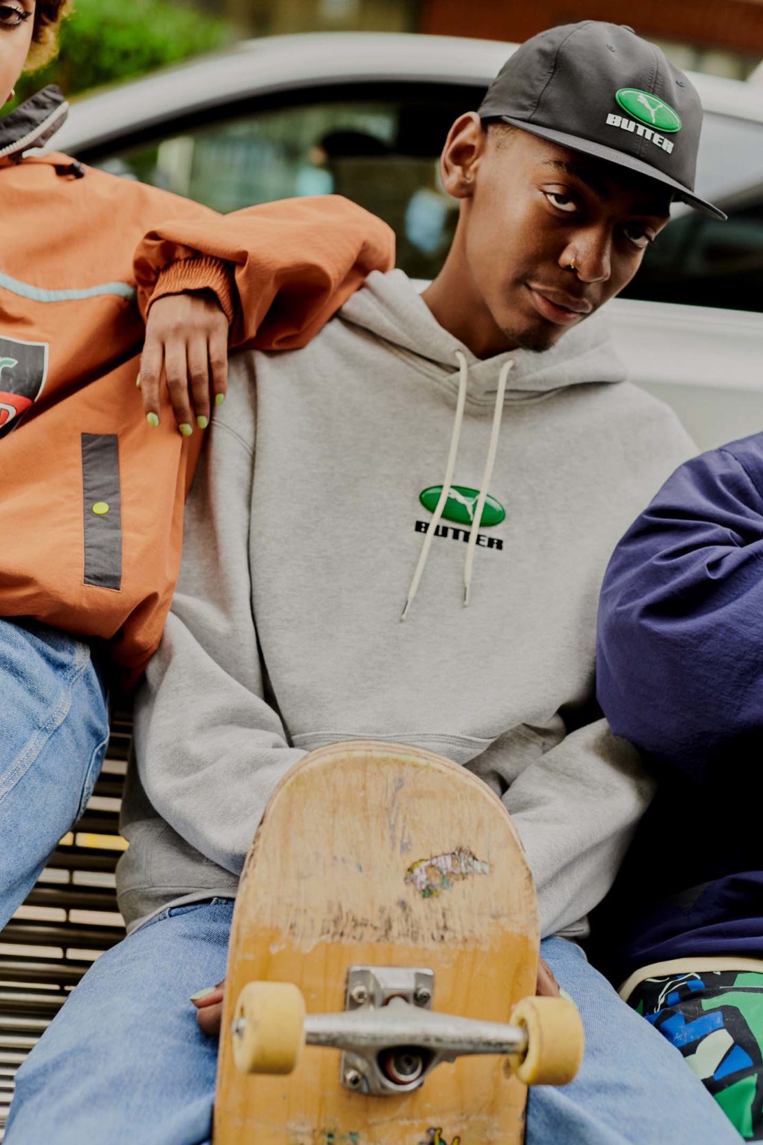 Teen in grey Butter Goods hoodie poses with a skateboard, baseball cap, and other people for a promotional fashion shot outdoors.
