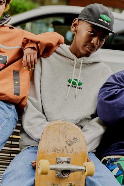 Teen in grey Butter Goods hoodie poses with a skateboard, baseball cap, and other people for a promotional fashion shot outdoors.