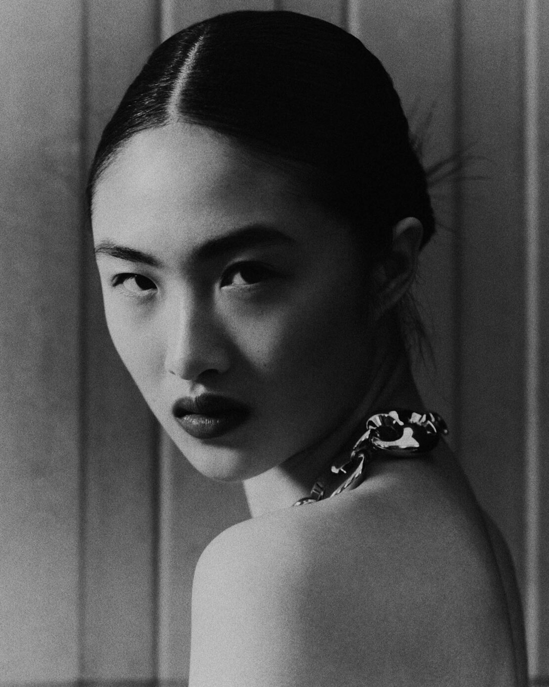Striking black-and-white portrait of an Asian woman with dark lipstick, wearing a chain necklace and gazing over her shoulder.