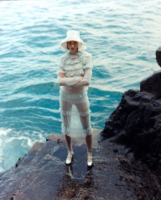 Fashion shot of a model wearing a ruffled dress and hat on a rocky shore with the sea in the background. Arms crossed and fair skin.