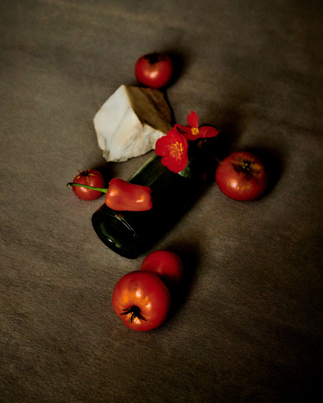 Still life photograph of a black bottle surrounded by red tomatoes, a red pepper, red flowers, and a piece of white marble on a gray background.