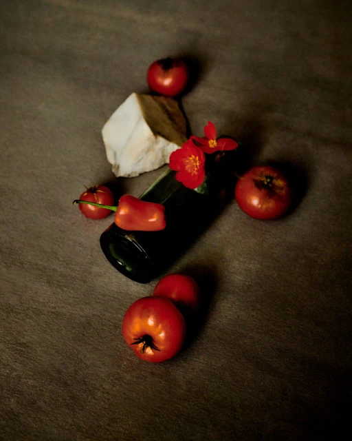 Still life photograph of a black bottle surrounded by red tomatoes, a red pepper, red flowers, and a piece of white marble on a gray background.