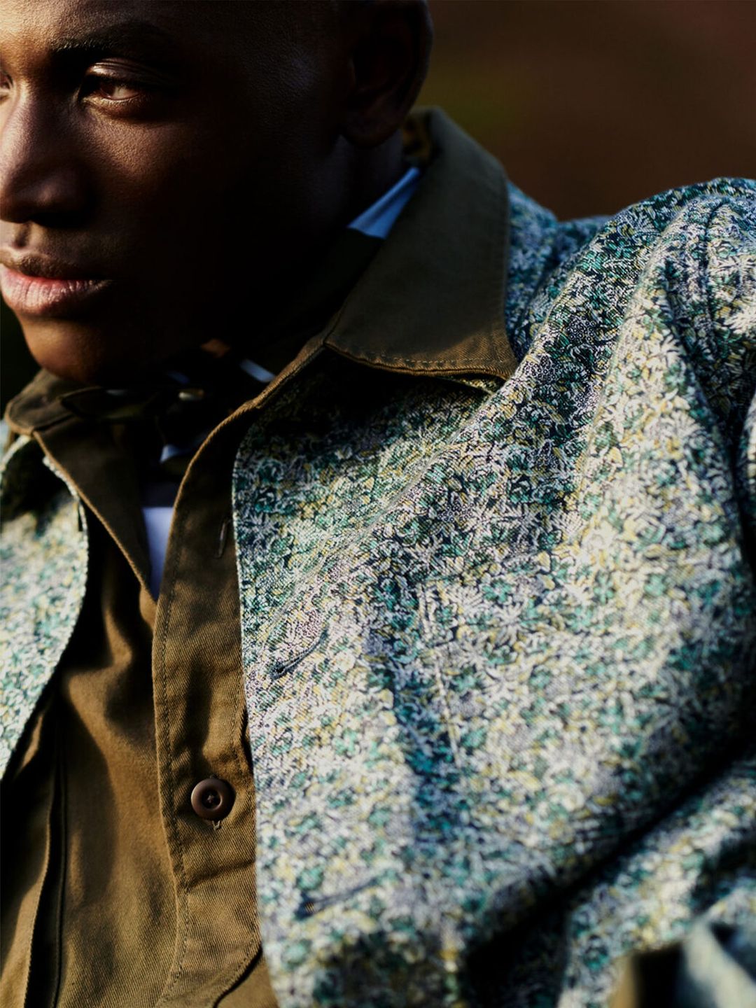 Close-up of a dark-skinned male wearing a button-down jacket with a unique pattern and an olive-green collared shirt outdoors.