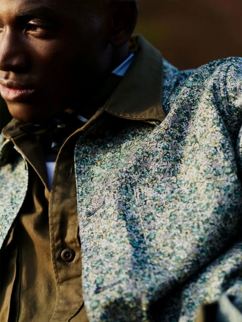 Close-up of a dark-skinned male wearing a button-down jacket with a unique pattern and an olive-green collared shirt outdoors.