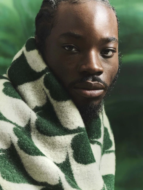 Close-up portrait of a Black man wrapped in a green and white patterned blanket, with a blurred green background. Fashion and style.