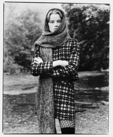 Black and white portrait of a woman wearing a plaid coat and a matching headscarf with arms crossed outdoors.
