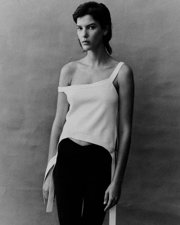 Portrait of a woman wearing an asymmetrical white top and dark trousers against a textured grey background by Daniel Benson.