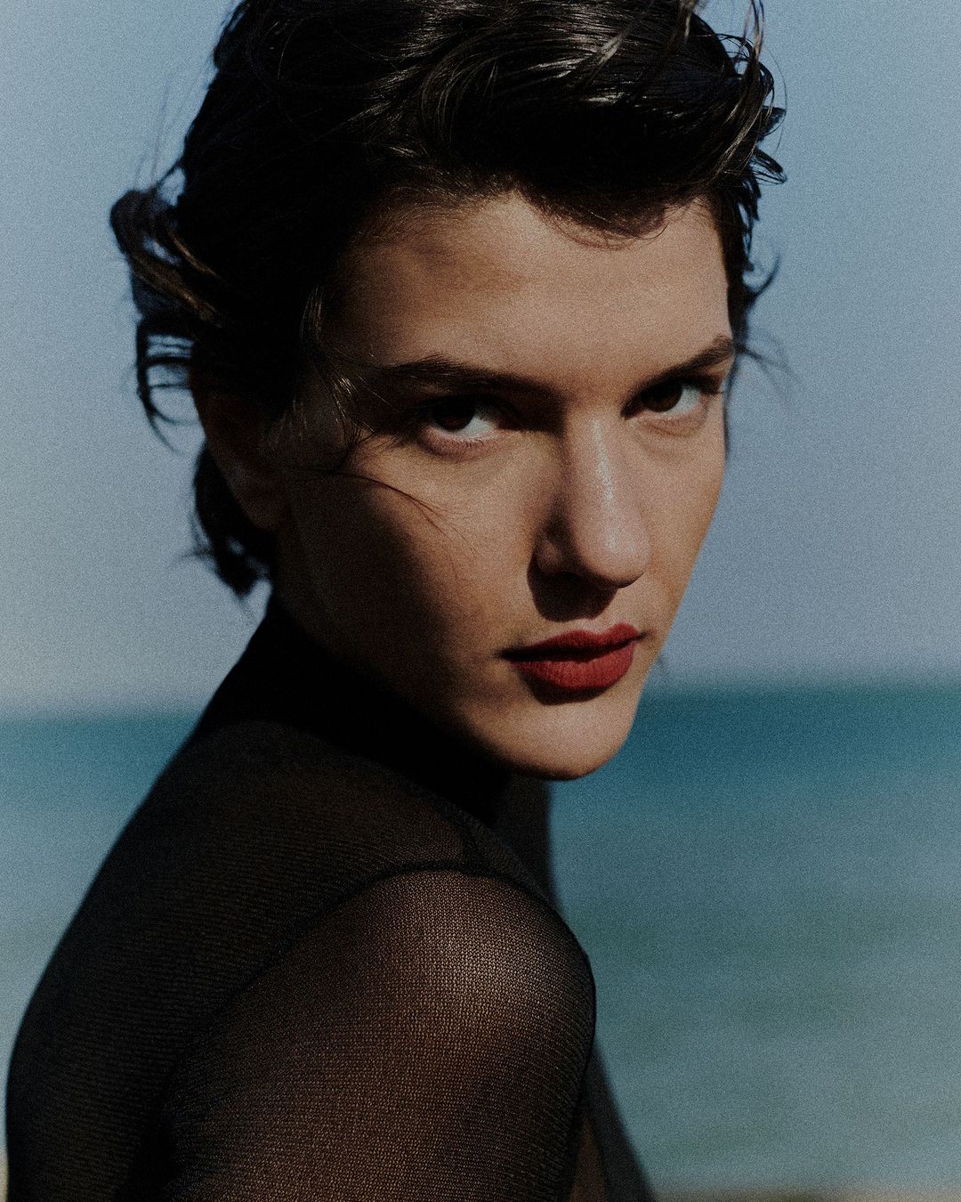 Portrait of a brunette woman with red lipstick and sheer black top against a blurred blue sea background, capturing a sophisticated and stylish look.