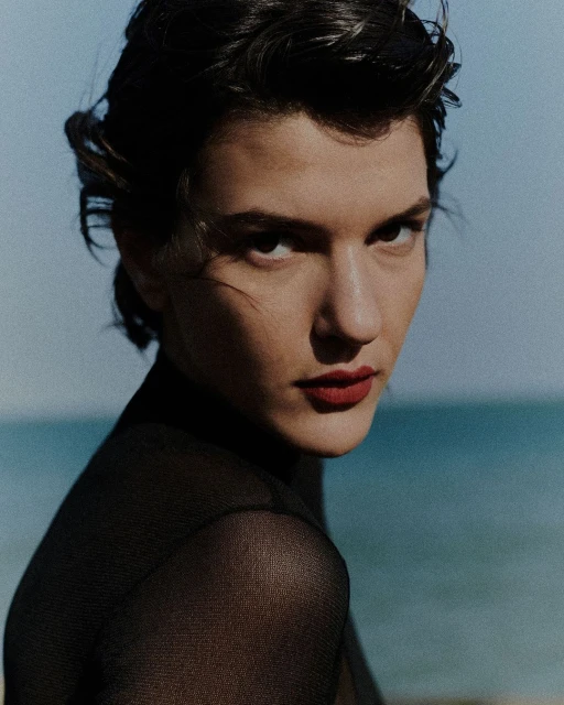 Portrait of a brunette woman with red lipstick and sheer black top against a blurred blue sea background, capturing a sophisticated and stylish look.