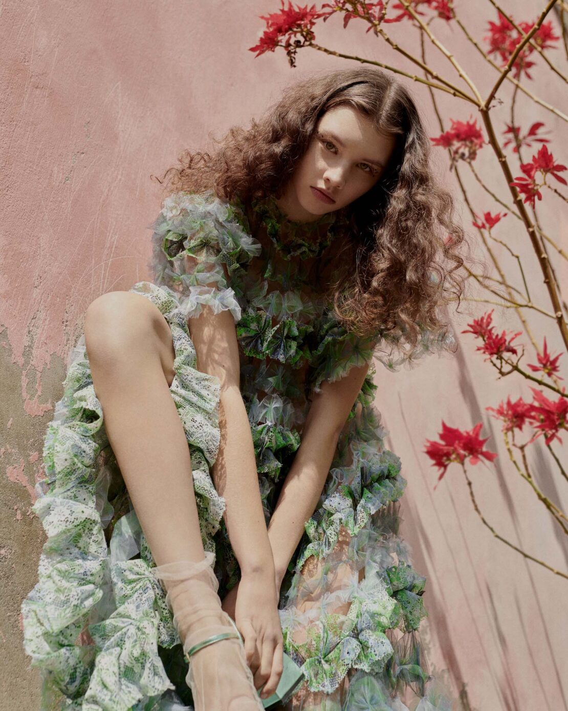 Young woman poses in a floral dress against a pink wall with red flowers, showcasing fashion, femininity, and a light-hearted aesthetic.