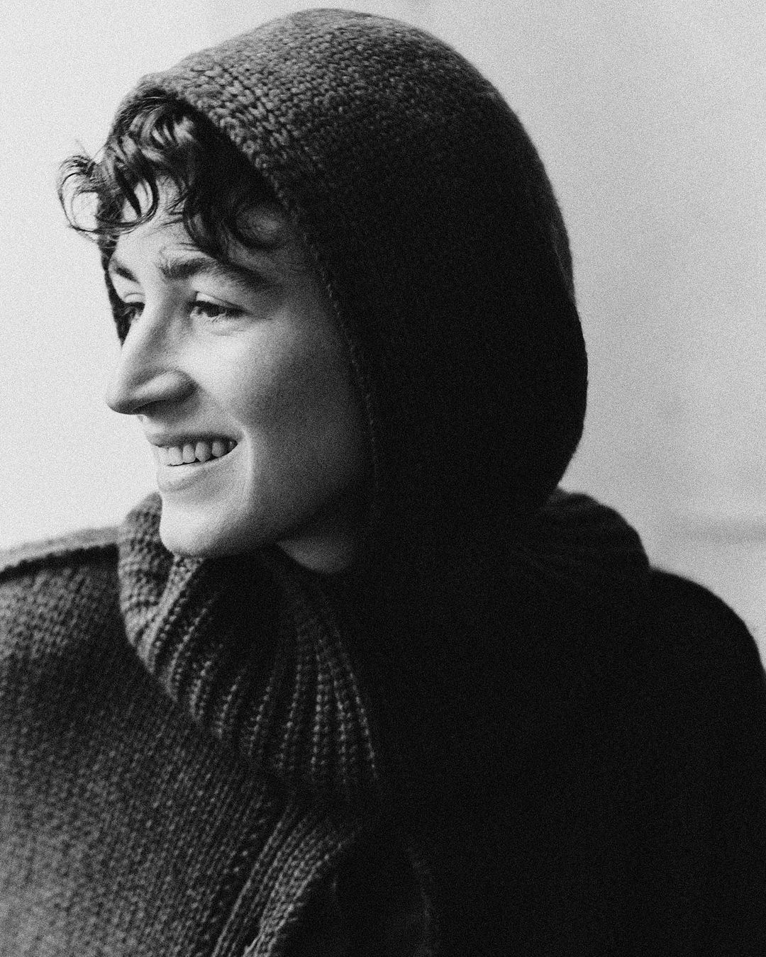 Close-up portrait of a smiling adult wearing a hooded sweater, featuring a monochromatic rendering, textured knitwear, and soft lighting.