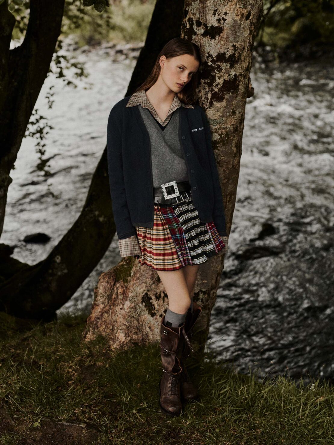 A young woman in a plaid skirt and boots leans against a tree near a flowing river in an outdoor fashion shot.