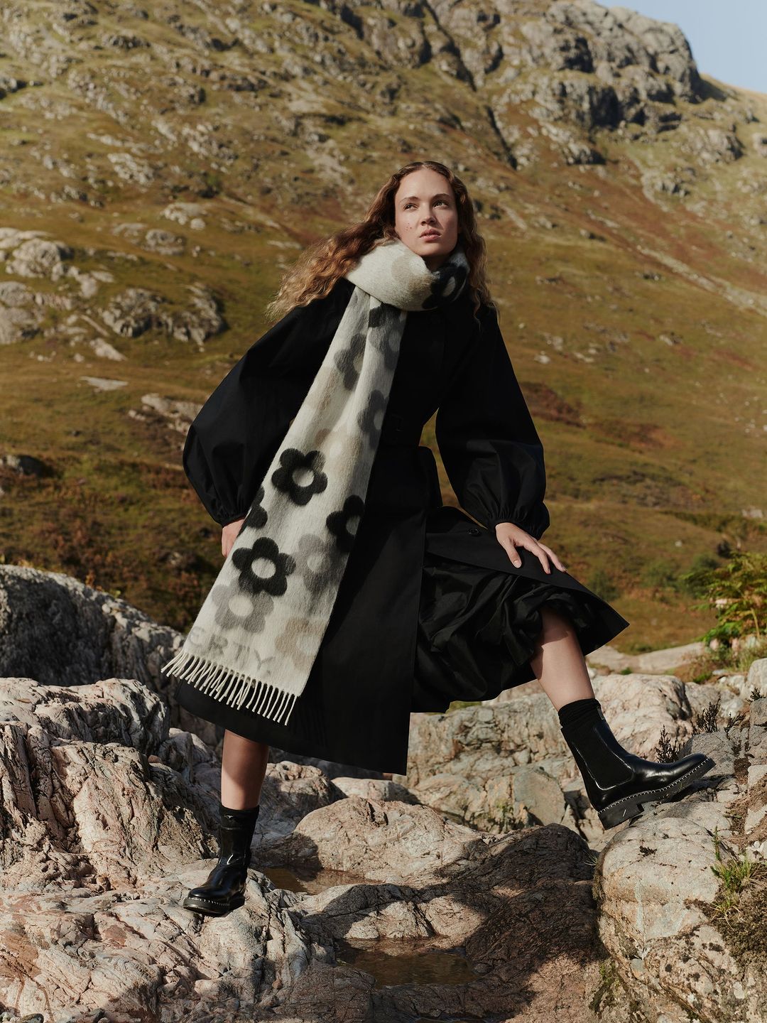 Woman in black dress and floral scarf posing on rocks with mountains in background, fashion photography, daytime outdoor setting.