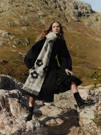 Woman in black dress and floral scarf posing on rocks with mountains in background, fashion photography, daytime outdoor setting.