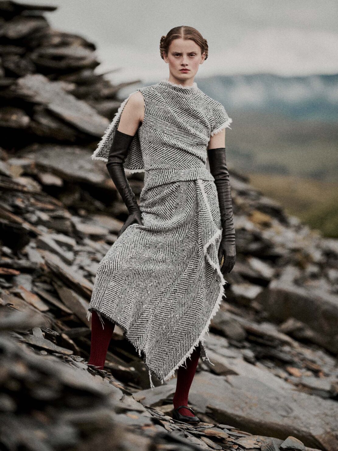 A female fashion model wearing a gray dress with arm gloves and red tights poses on rocky terrain against a blurred landscape background.