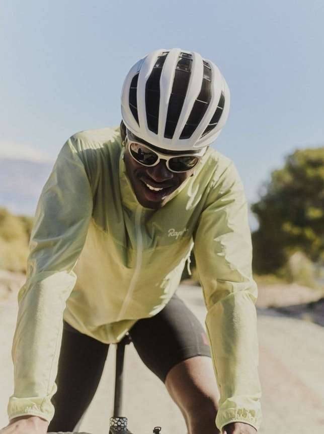 Cyclist smiles while riding on a paved road in the sunlight wearing a helmet, sunglasses, and a yellow jacket in an outdoor setting.