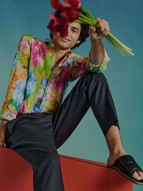 Adult male seated on a red chair against a light blue backdrop, wearing a colorful floral shirt, dark pants, slides, and holding red flowers.