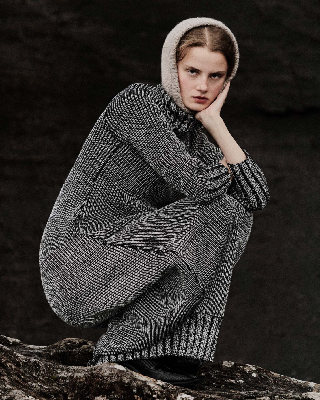 A woman in a knitted dress and hat sits on a rock in a studio shot, looking at the camera with a neutral expression in dim lighting.