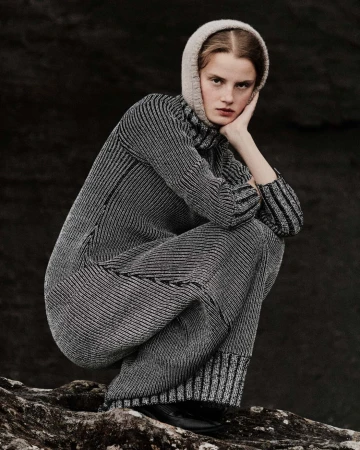 A woman in a knitted dress and hat sits on a rock in a studio shot, looking at the camera with a neutral expression in dim lighting.