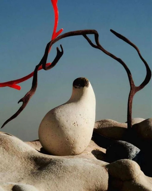 Stone sculpture with abstract branch-like objects in the foreground set against a clear blue sky in an outdoor environment.