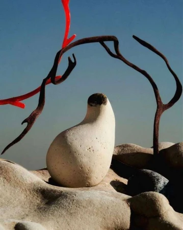Stone sculpture with abstract branch-like objects in the foreground set against a clear blue sky in an outdoor environment.